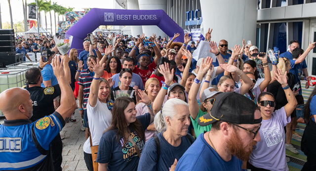 Large crowd of participants walking under a purple Best Buddies arch at an outdoor event, with many wearing event t-shirts.